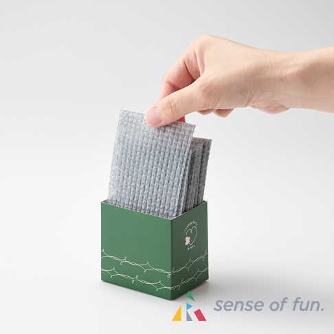 一疊淺灰色、帶有鬆餅紋路的潔淨海綿巾，被置於綠色紙盒中，一隻手正在從中抽取一張。