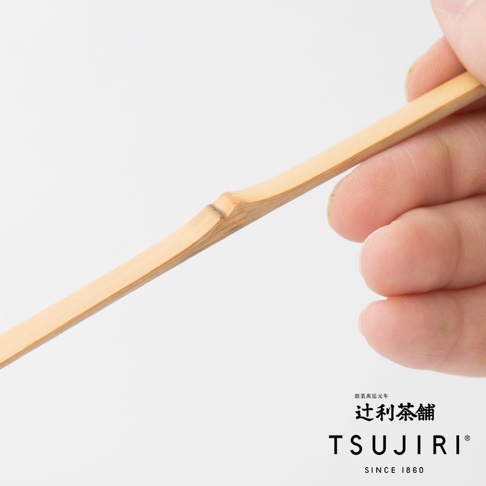 一雙手正拿著一根淺棕色、質地天然的竹製抹茶杓，杓柄上有明顯的竹節紋理。