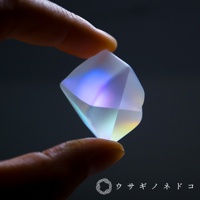 手指夾著一個無色透明的稜鏡石，稜鏡石表面呈現出藍色、紫色、黃色和綠色的光暈，材質為玻璃或水晶，具有多個切割面。