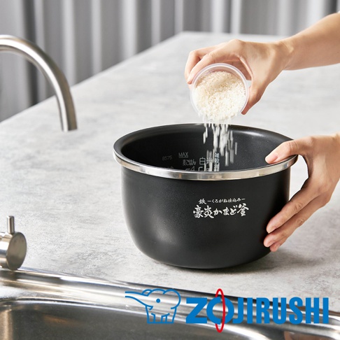 黑色金屬內鍋，內側有水位刻度，外側印有日文品牌標誌。一隻手正將米從透明容器中倒入內鍋。