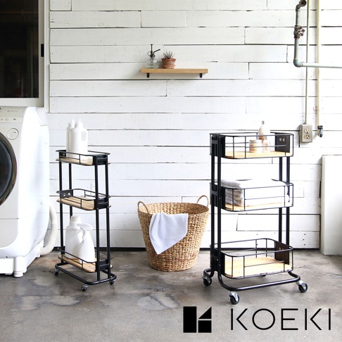 黑色金屬三層置物夾縫式收納推車，配有木質層板，可移動，有兩個置物架，其中一個架子上有白色瓶狀物，另一個架子上有疊好的白色毛巾。