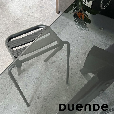 一張黑色金屬吧檯椅，椅面為條狀紋路，椅腳呈彎曲狀，置於玻璃桌旁，背景為灰色地磚。