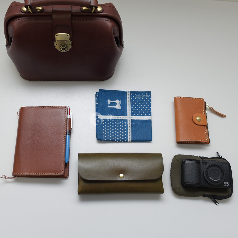 深棕色皮革迷你醫生包，附黃銅鎖扣。旁邊擺放著一個棕色皮革筆記本、藍色印花方巾、棕色皮革卡夾、綠色皮革長夾，以及一個裝有黑色數位相機的保護套。
