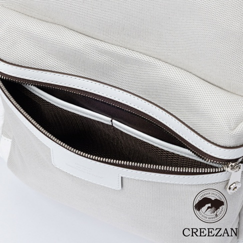 這款 CREEZAN 白色後背包的特寫鏡頭，展示了其精緻的質感。主體由高品質織紋帆布製成，搭配白色皮革飾邊與深棕色內襯，前置拉鍊口袋處刻有 CREEZAN 品牌標誌與手工製造產地資訊，整體設計簡約且做工細膩。