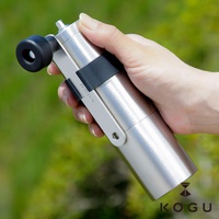 手持一個銀色圓柱形日製18-8不鏽鋼手搖咖啡磨豆器，帶有黑色旋鈕和黑色束帶，背景為模糊的綠色植物。