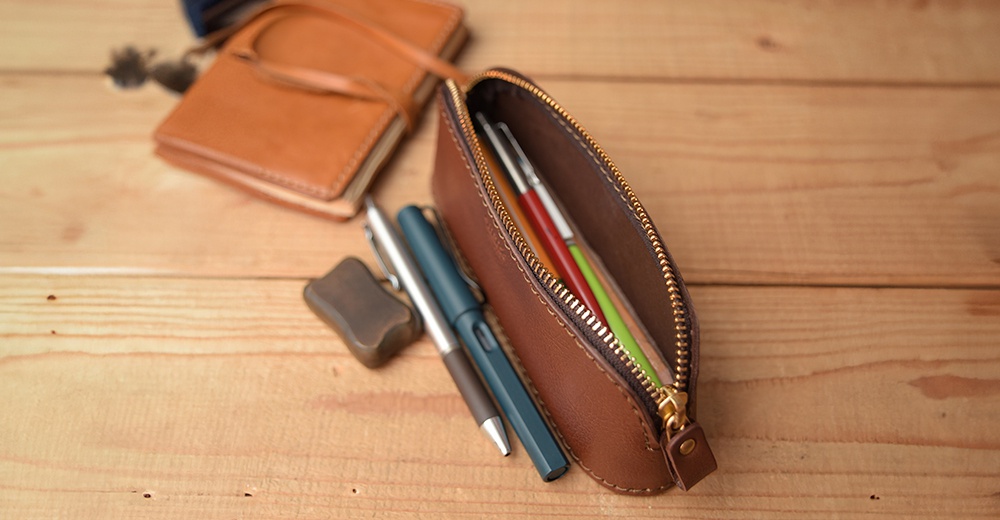 職人手作皮革三角弧形收納筆袋，棕色皮革，金色拉鍊，內有綠、紅、銀色筆。