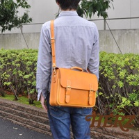 職人手作的駝色皮革三用公事包，帶有雙扣環和提把，斜背於穿著藍色襯衫和牛仔褲的男士身上。