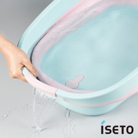 淺藍色和粉色搭配的伸縮浴盆，內部有水，一隻手正提起浴盆邊緣，水從浴盆中傾倒出來，顯示其靈活的結構。