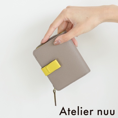一隻手正輕巧地拿著一個 Atelier nuu 品牌推出的灰色皮革短夾，短夾表面呈現細緻的荔枝紋路，中間配有鮮明的黃色皮革飾帶作為扣飾，側邊則附有拉鍊拉環，整體設計簡約且充滿質感。