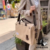 米色純綿帆布製成的商務公事包，配有棕色皮革提把和可拆卸肩帶，包款正面有一個大型翻蓋口袋，側邊有皮革飾件，適合 B4 尺寸文件。