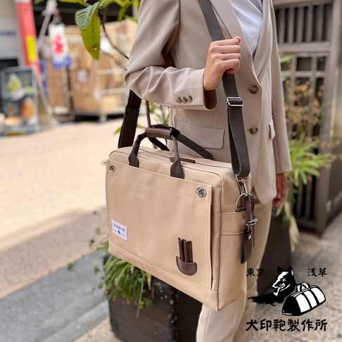 米色純綿帆布製成的商務公事包，配有棕色皮革提把和可拆卸肩帶，包款正面有一個大型翻蓋口袋，側邊有皮革飾件，適合 B4 尺寸文件。