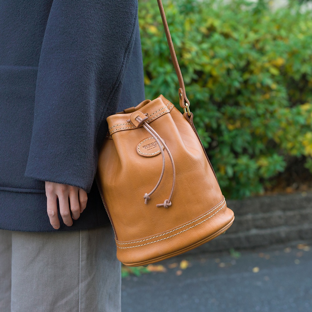 圓筒形棕色皮革束口單肩包，配有細長肩帶和束繩。包身車線明顯，皮革壓印品牌標誌。