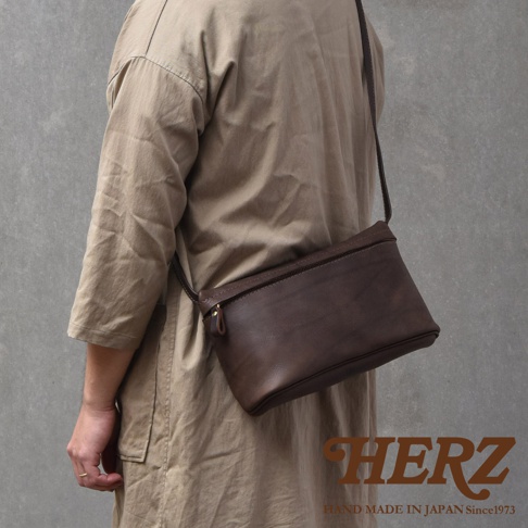 深棕色皮革製成的斜背包，由人斜背於身，包款有拉鍊開口，右下角有HERZ字樣及HAND MADE IN JAPAN Since1973字樣