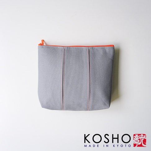 京都KOSHO帆布化妝收納包L，整體為簡約灰色帆布材質，包身設有兩道細緻的垂直拼接縫線設計，頂部配有亮眼的橘色拉鍊，呈現時尚質感，右下角印有KOSHO MADE IN KYOTO品牌標誌與紅色印章字樣。