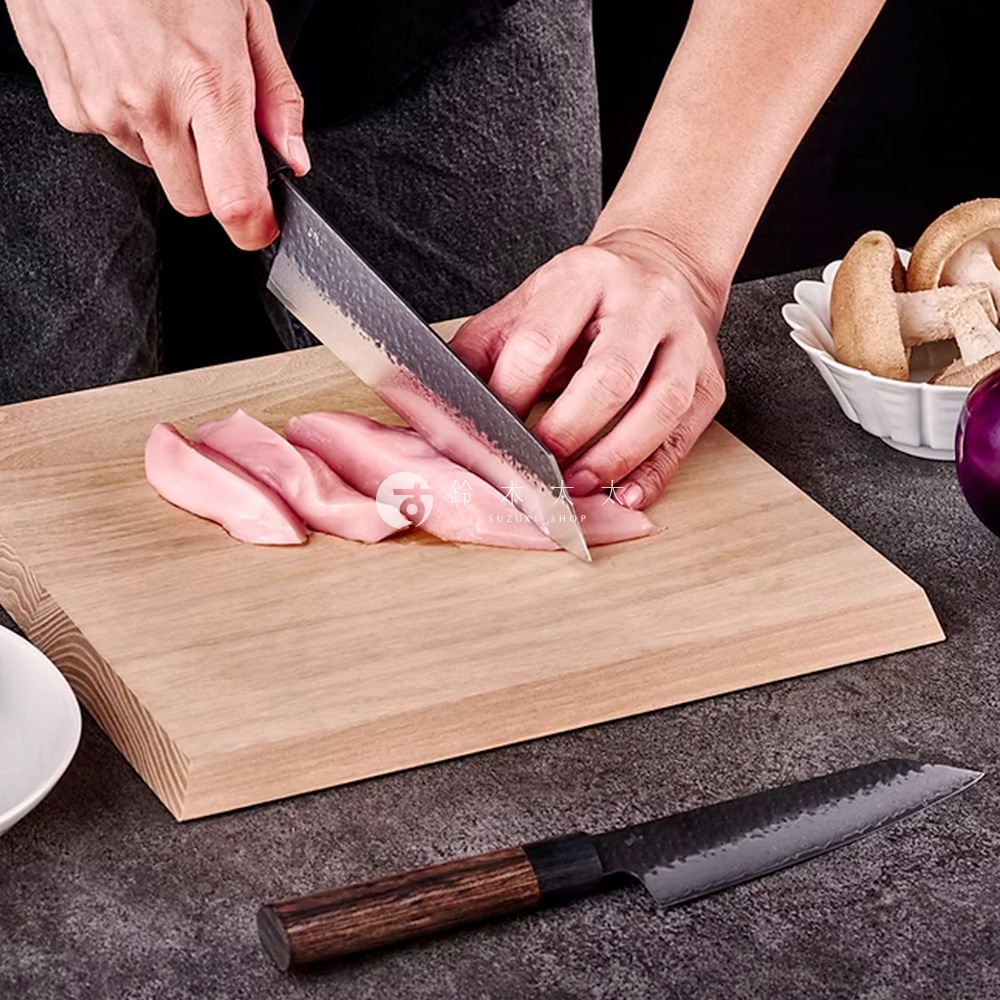 玄 / Bunka 文化刀19cm，刀身為銀色不鏽鋼，帶有鎚紋，黑色木質刀柄。刀正被用於切割粉紅色肉類，置於淺木色砧板上。背景有香菇和紫色洋蔥。