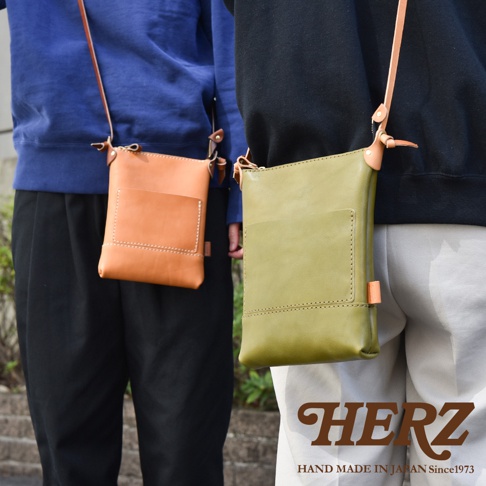 兩款職人手作皮革直式隨身單肩包，分別為焦糖色和橄欖綠，皆為簡約的長方形設計，配有同色系或淺棕色皮革的細長肩帶。包身正面設有一個較淺的貼袋，由皮革邊緣縫線和包身縫線清晰可見。