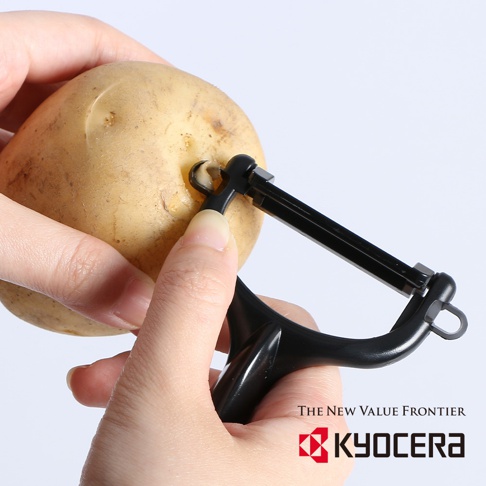 一只手拿著一顆帶有泥土的馬鈴薯，另一隻手使用黑色陶瓷削皮刀的刀刃去除馬鈴薯皮。