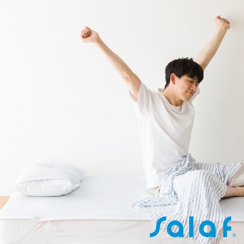 白色床墊上有一個淺藍色枕頭，一位穿著白色T恤的男子坐在床墊上伸展身體，身上蓋著一條藍白條紋的毛巾。