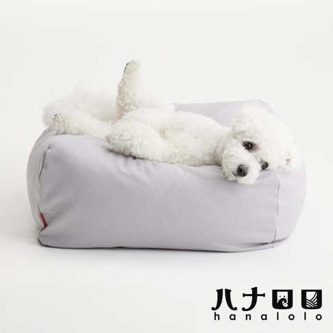 一隻白色比熊犬舒適地趴在灰色的方形懶骨頭寵物沙發上，狗狗姿勢放鬆，右後腿微微翹起，頭部靠在沙發邊緣，畫面右下角有日文品牌標識hanalolo。