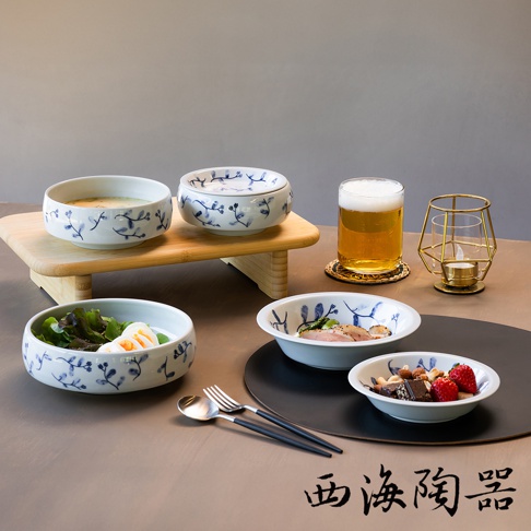 波佐見燒藍花紋六件式碗盤組，包含多個白色陶瓷碗盤，皆繪有藍色植物花紋。其中一個碗盤內盛有沙拉和水煮蛋，另一個碗盤內有堅果、巧克力和草莓。畫面右側擺放著一杯啤酒和一個金色金屬燭台。