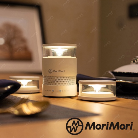 松本素白LASMO Speaker燭光氣氛音響，圓柱形主體，頂部為透明燈罩，內有LED燈，底部為網狀喇叭，側面有MoriMori品牌標誌及控制按鈕。