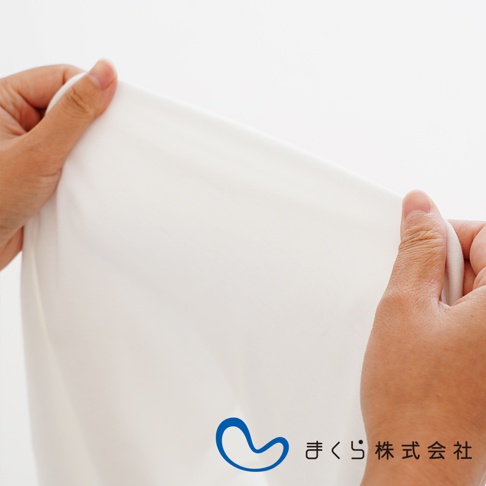 雙手撐開白色柔軟的枕套，展示其材質的細緻紋理，左側有藍色品牌標誌及日文公司名稱。