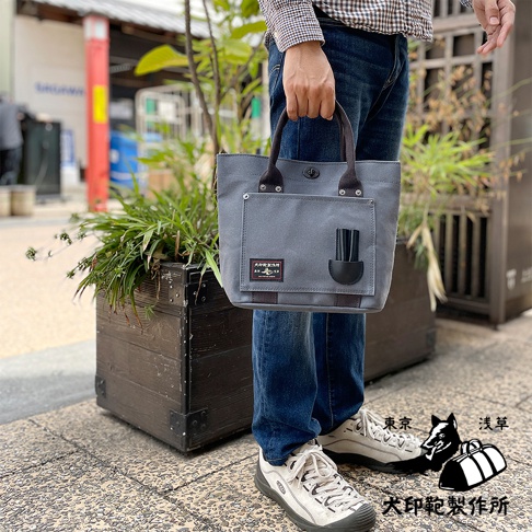 灰色帆布水桶托特包，配有深棕色提把和黑色皮革裝飾，正面有一個方形口袋，內有兩個黑色筆狀物品。