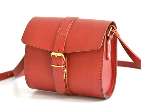 紅色皮革製成的單肩包，包蓋上有皮扣和黃銅搭扣，肩帶亦由皮革製成，縫線明顯。