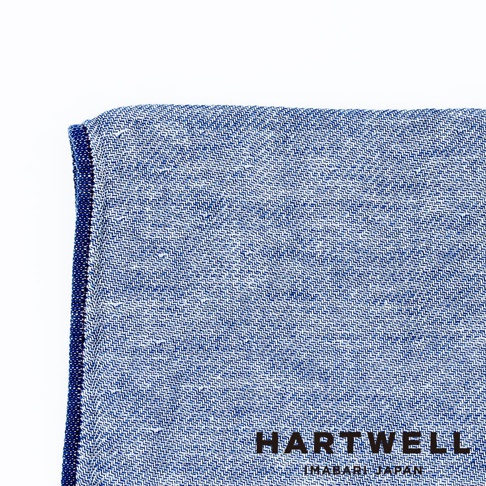 今治復古丹寧風雙織柔感毛巾，以藍色與白色交織的細緻織紋呈現，毛巾邊緣縫有深藍色滾邊，右下角印有黑色「HARTWELL」及「IMABARI JAPAN」字樣。
