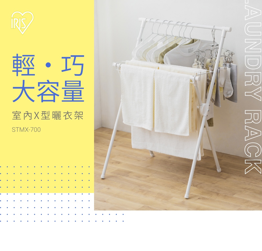 白色IRIS OHYAMA品牌室內X型曬衣架STMX-700，放置於室內木質地板上，頂部掛桿掛有數件衣物，下方架層則晾曬白色毛巾，整體設計簡約輕巧且具備多層晾曬功能，呈現出居家收納與衣物清潔的視覺情境。