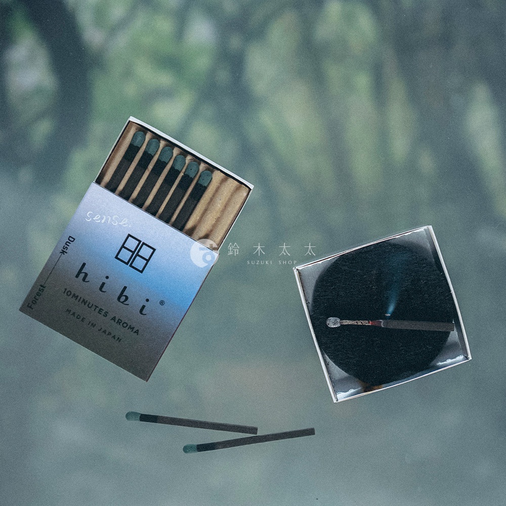 這款 hibi 香氛火柴禮盒以俯視角度呈現，畫面左側為半開的火柴盒，露出數支帶有深綠色火柴頭的香氛棒；右側為放置在黑色圓形底座上正燃燒冒煙的火柴，下方則有兩支平放的火柴，整體呈現簡約精緻的質感。