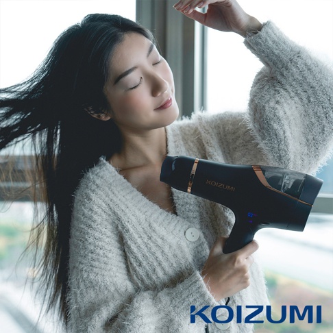 一位女性正使用黑色、帶有玫瑰金飾邊的 KOIZUMI 負離子吹風機，吹整她的長髮。吹風機機身有一塊顯示屏，顯示數字「03」。