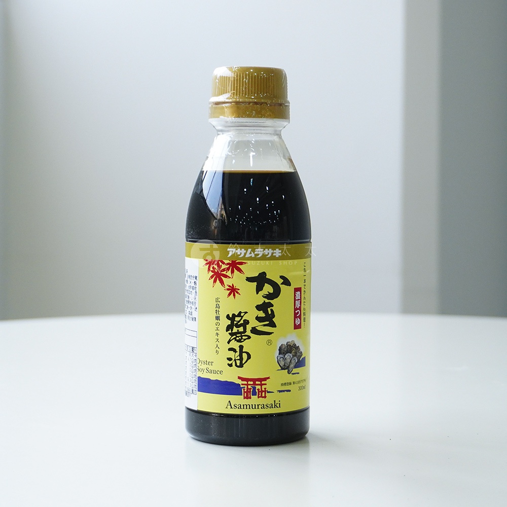 一瓶放在白色桌面上的朝紫廣島牡蠣醬油，瓶身為透明材質裝有深色液體，並蓋有金色瓶蓋。產品標籤以黃色為底，印有紅色楓葉圖案、牡蠣插畫以及紅色鳥居圖示，上方明確標示品牌名稱Asamurasaki與商品名稱かき醤油，瓶身正中央印有醒目的黑色漢字。