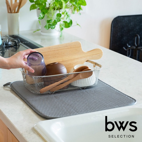 灰色可折疊收納瀝水墊上放置著一個銀色金屬網籃，籃內裝有紫色玻璃杯、木質砧板、棕色碗狀物、白色小碗和木勺。