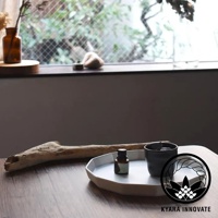 KYARA INNOVATE 品牌 logo，旁有一小瓶精油、黑色杯子和一個樹脂擴香石，擺放在一個低矮的盤子上，後面是飄窗和植被。