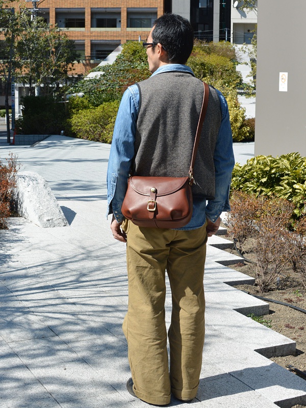男士肩背棕色皮革馬鞍包，包身圓潤，帶有金屬扣環，搭配牛仔襯衫、針織背心與卡其色長褲。