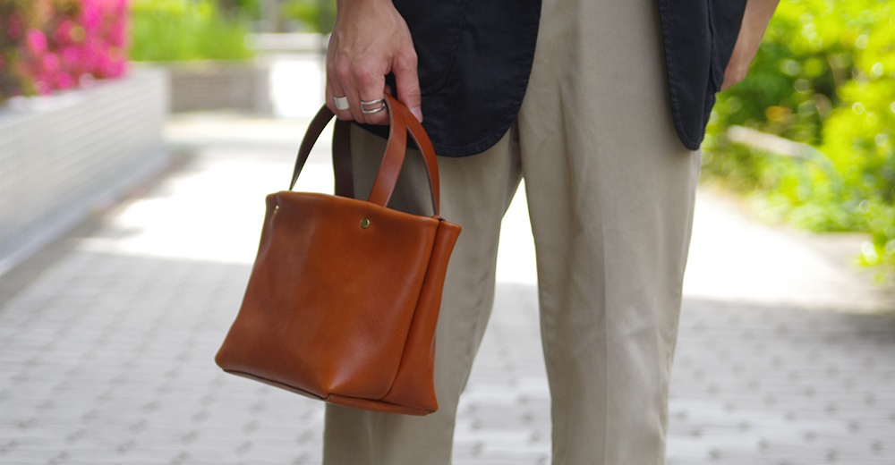 職人手作的棕色皮革托特包，配有兩個短提把，包身簡潔，正面有一個黃銅鈕扣。