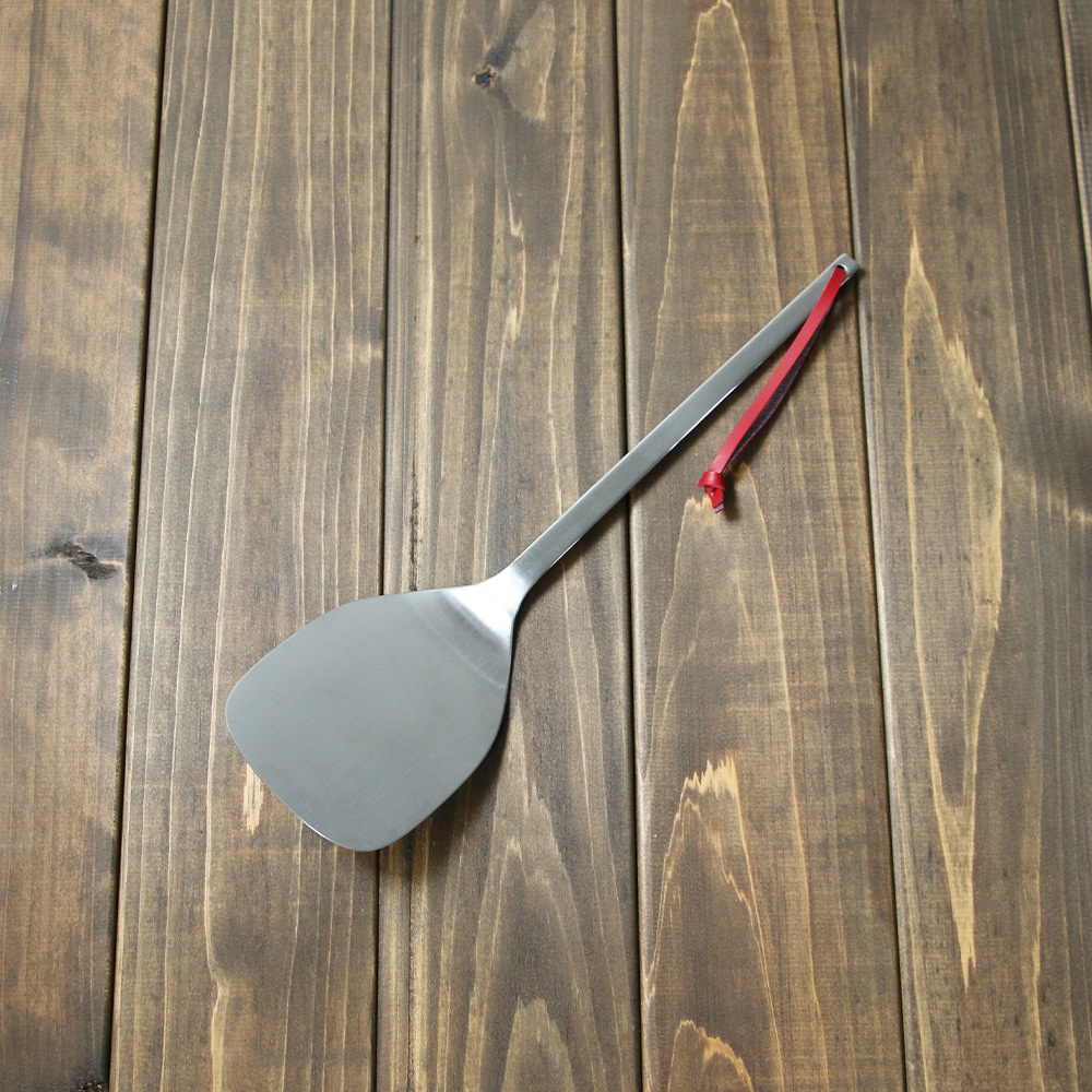 銀色不鏽鋼萬用鍋鏟，帶有紅色掛繩，置於深棕色木紋桌面。