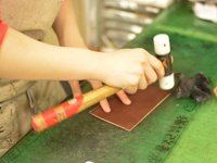 職人手作皮革存摺文件收納夾 (KZ-144)，使用棕色皮革製作，由師傅手持木柄鐵鎚和圓形壓具，在綠色工作台上進行加工。