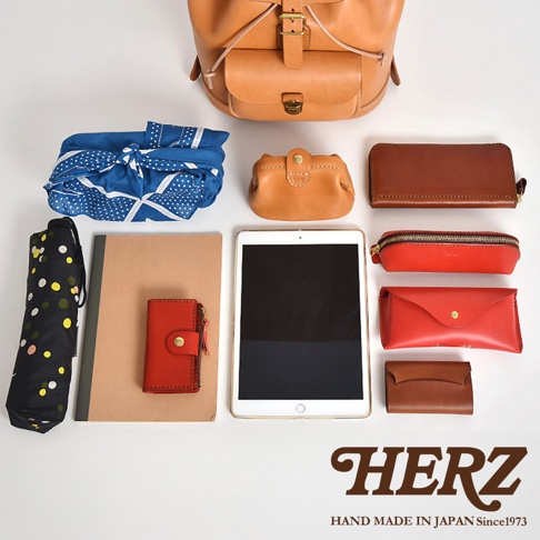 職人手作皮革經典後背包 R-4，搭配各式皮革配件，包含：小零錢包、長夾、筆袋、眼鏡盒、卡夾，以及藍色波點包巾、黑色圓點摺疊傘和白色邊框平板電腦，展現多樣化的收納組合。