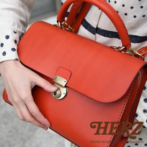 日本HERZ職人手作紅色皮革多功能收納單肩包，特寫包包的金屬鎖頭細節，下方可見HERZ品牌標誌及Hand made in Japan Since1973字樣