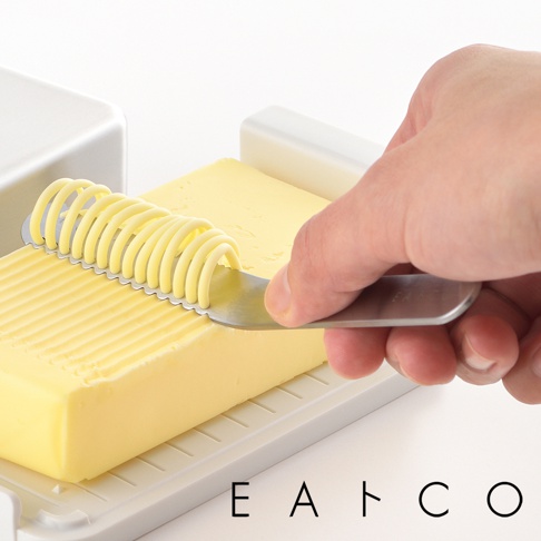 日製奶油刀，不鏽鋼材質，刀刃帶有梳狀紋路，正將黃色奶油刮出螺旋狀。奶油置於白色奶油盒中，盒底有條紋。