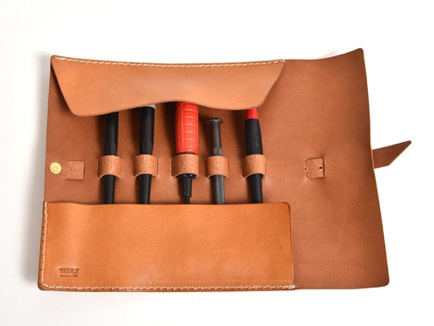 淺棕色皮革捲捲收納筆袋，內有四支筆，其中一支為紅色握把，袋口有金屬壓扣，皮革上印有品牌標誌。