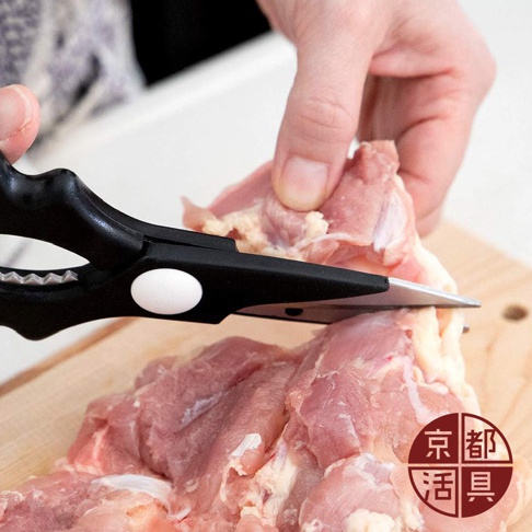 一雙黑色手柄的不鏽鋼萬用料理剪刀，正在切割一塊生雞肉，剪刀刀刃鋒利，手柄上有白色圓形鉚釘，背景為木質砧板。