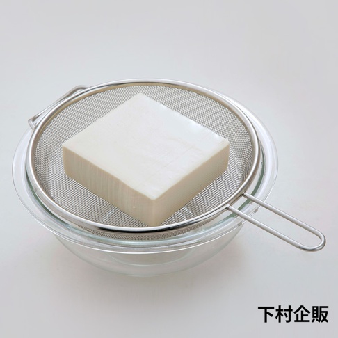 不鏽鋼濾網內放著一塊白色豆腐，濾網置於玻璃碗上方，濾網邊緣和手柄為銀色不鏽鋼材質，網孔細密。
