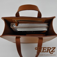 職人手作皮革直式托特包，淺棕色皮革，包內裝有筆記本和畫冊，頂部有拉鍊隔層和提把。