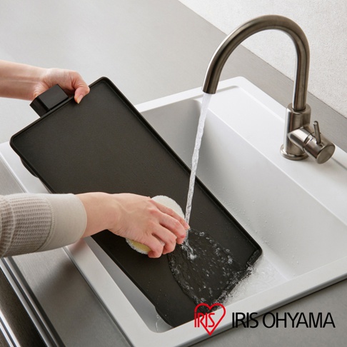 雙手正拿著黑色輕薄電烤盤，在水龍頭下沖洗。一隻手握住烤盤的黑色手柄，另一隻手拿著白色海綿擦拭烤盤表面，水流從水龍頭流下。