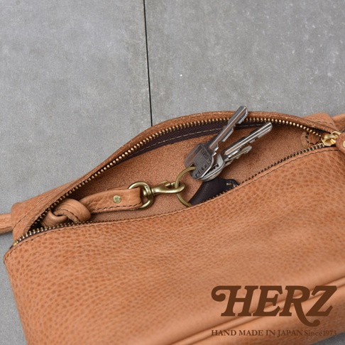 HERZ 品牌標誌，下方印有 HAND MADE IN JAPAN Since1973 字樣，一條棕色皮革斜背包的拉鍊打開，露出內側，並掛有鑰匙串。