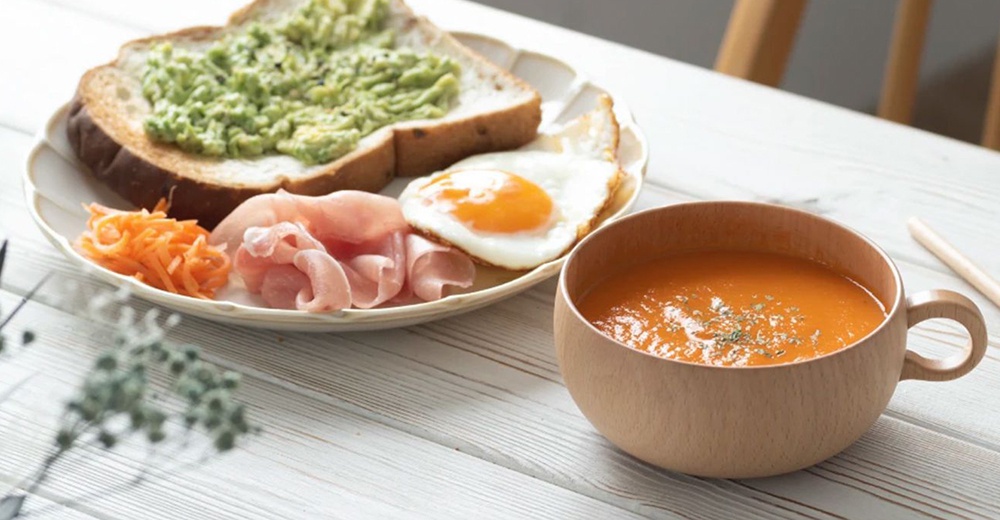 楓木湯杯內盛有橘紅色湯品，湯品表面撒有少量綠色香料。湯杯為圓形，木紋清晰，帶有木質把手。