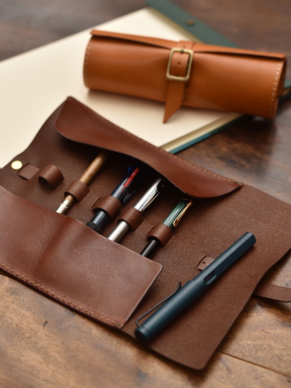職人手作的棕色皮革捲捲收納筆袋，內部有四個筆槽，可固定鋼筆、原子筆等書寫工具，外部則有一個金屬釦帶。